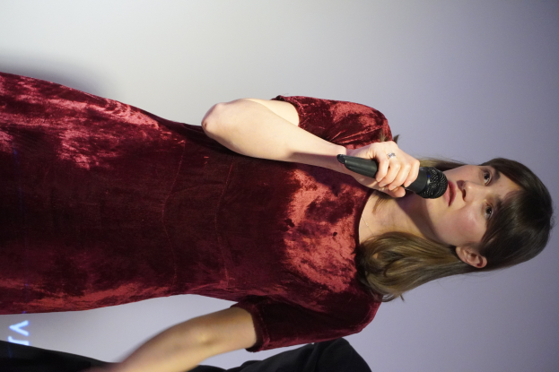 Eine Frau in einem roten Kleid steht selbstbewusst auf einer Bühne, hält ein Mikrofon und hat einen eleganten Bob-Schnitt und natürliches Make-up.