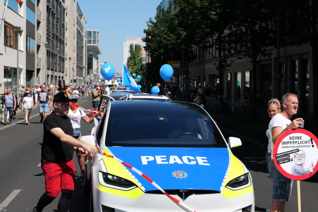 Eine Gruppe von Menschen geht neben einem Polizeiwagen her, der mit blauen, gelben und roten Schleifen geschmückt ist und auf dem das Wort "Frieden" zu sehen ist, vor einem Hintergrund aus Gebäuden, Bäumen und Flaggen unter einem klaren blauen Himmel.