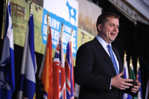 Mann in Anzug und Krawatte, der vor Mikrofonen auf einem Podium spricht, umgeben von Flaggen und einem Bildschirm im Hintergrund.