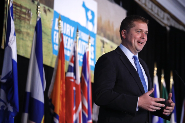 Mann in Anzug und Krawatte, der vor Mikrofonen auf einem Podium spricht, umgeben von Flaggen und einem Bildschirm im Hintergrund.