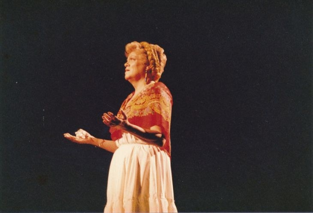 Eine Frau in einem weißen, hochgeschlossenen, langärmeligen Kleid steht auf einer Bühne mit ausgestreckten Armen, beleuchtet vor einem dunklen Hintergrund, mit einem Dutt und einem ernsten Gesichtsausdruck.