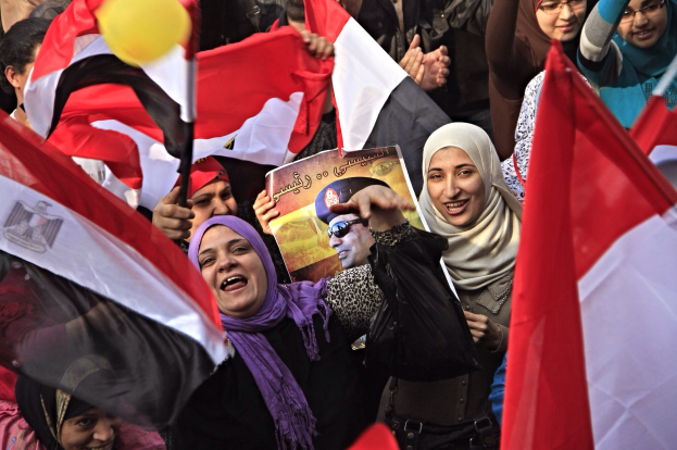 Eine Gruppe ägyptischer Demonstranten auf dem Tahrir-Platz in Kairo, die Fahnen schwenken und Parolen rufen, wobei einige Schals tragen und einer ein Schild hält.