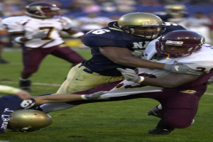 Zwei Fußballspieler in Helmen und Uniformen kollidieren auf dem Feld, während andere im Hintergrund zuschauen.