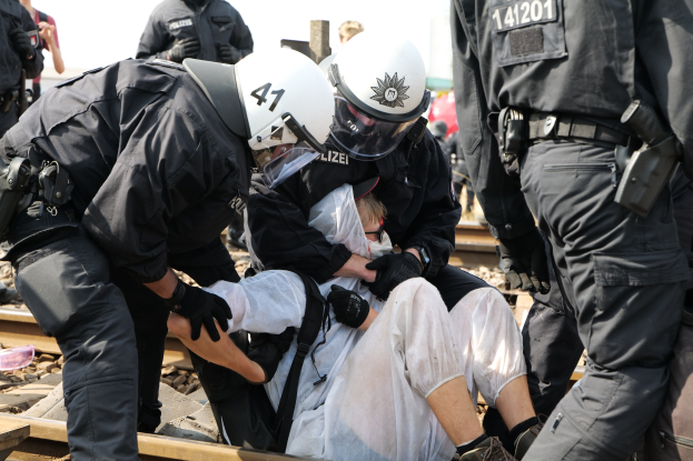 Eine Gruppe von Polizisten in Einsatzausrüstung nimmt einen Mann in einem weißen Kleid auf einem Bahngleis fest, während andere im Hintergrund stehen.