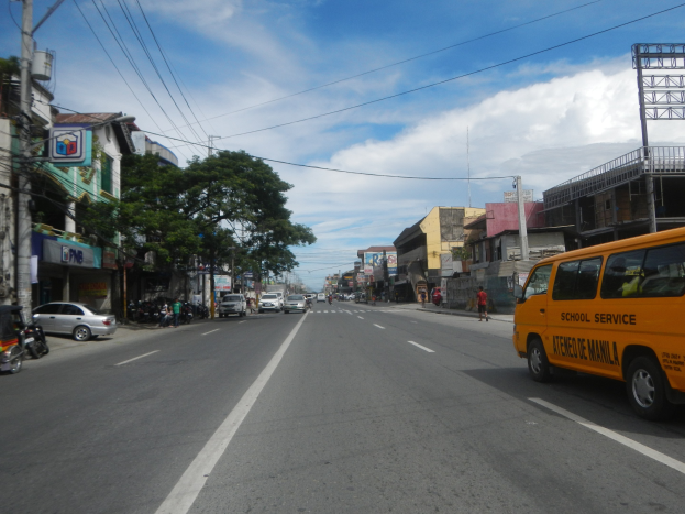Ein gelber Schulbus fährt auf einer Straße mit hohen Gebäuden, Passanten, Bäumen, Strommasten und Texttafeln unter einem bewölkten Himmel.