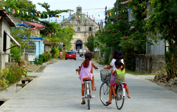 Drei Kinder fahren Fahrräder auf einer von Bäumen gesäumten Straße mit Gebäuden auf beiden Seiten, einem Auto und einem Gebäude im Hintergrund und Himmel oben