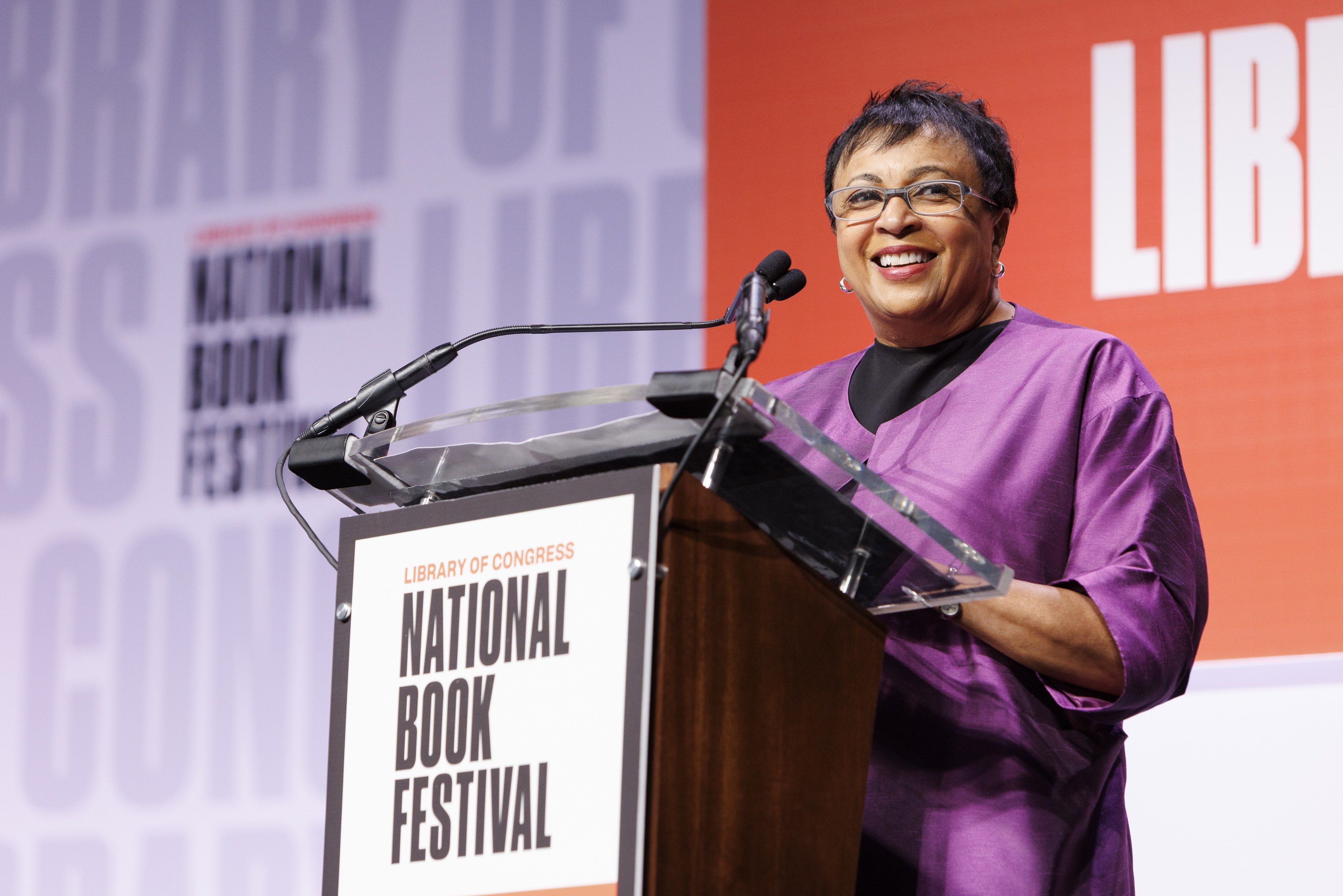 Eine Frau in einem purpurfarbenen Kleid steht an einem Podium mit zwei Mikrofonen und einem orange-weißen Banner mit der Aufschrift 'National Book Festival' im Hintergrund.