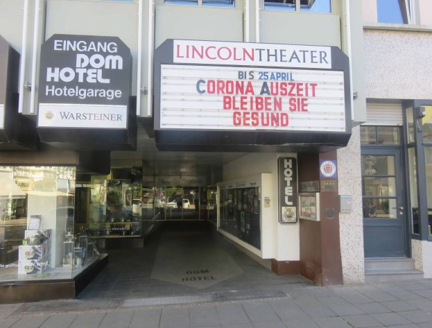 Außenansicht des Lincoln Theaters in Berlin, Deutschland, mit Glasfenstern und -türen sowie einer Tafel mit Text und einem Innenraum, der eine pulsierende Stadtlandschaft vermuten lässt.