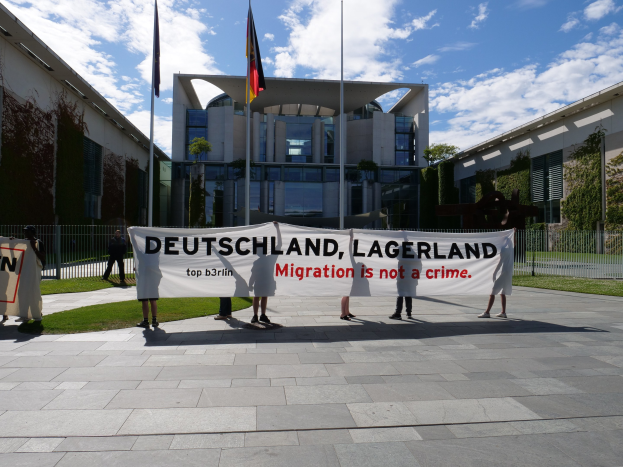 Eine Gruppe von Menschen mit einem Transparent mit der Aufschrift "Deutschland, Lagerland Migration ist kein Verbrechen" vor einem Gebäude mit Fenstern, umgeben von Gras, einem Metallzaun, Pflanzen, Bäumen und Fahnen auf Ständen unter einem bewölktem Himmel.