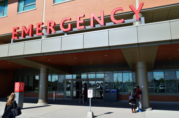 Gruppe von Menschen, die auf ein Notaufnahmeanbau mit Glaswänden, Säulen und einem sichtbaren Namensschild zugeht, begleitet von Straßenschildern und einem Müllcontainer.