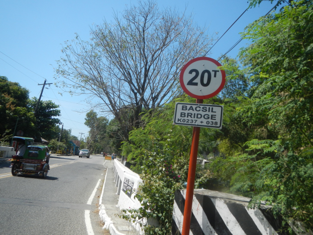 Eine Straße mit fahrenden Fahrzeugen, ein Geschwindigkeitsbegrenzungsschild, eine Trennwand, Bäume, Strommasten mit Drähten und einen klaren blauen Himmel im Hintergrund.
