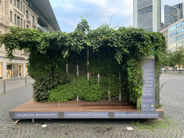 Ein grünes Gebäude mit Efeu an der Seite, umgeben von Bäumen, Fahrzeugen, Strommasten, Schildern und anderen Gebäuden unter einem bewölkten Himmel, Teil des Staufbilter Biodiversität nachhaltigen Gebäudes in Berlin, Deutschland.