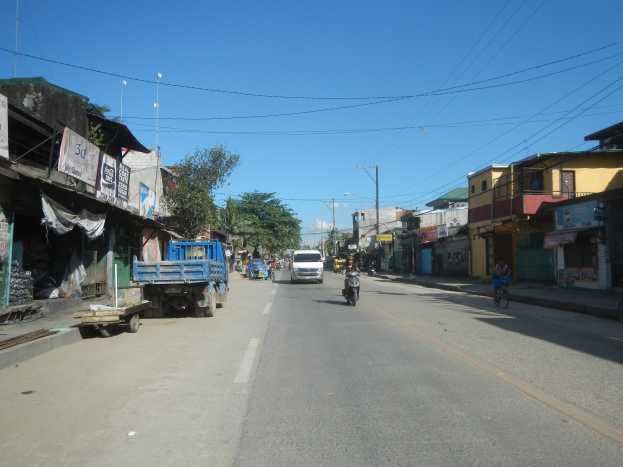Eine Straße in einer Stadt mit fahrenden Fahrzeugen, ein Radfahrer auf der rechten Seite, Gebäude auf beiden Seiten, Bäume im Hintergrund und ein klarer blauer Himmel.