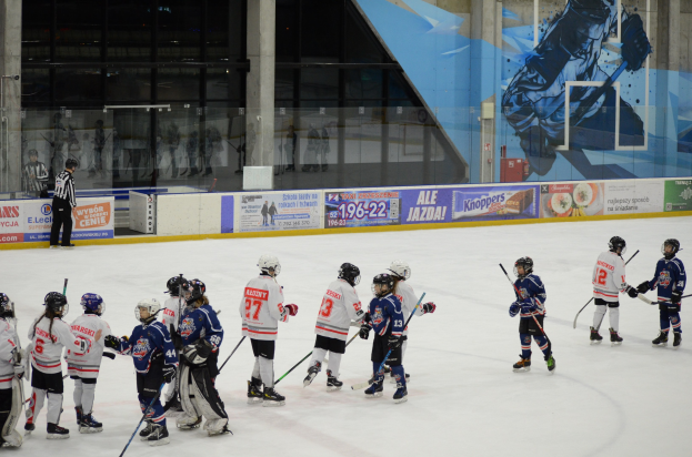 Gruppe junger Menschen, die Eis hockey auf einer Indoor-Eishalle spielen, mit Helmen und Stöcken, sowie einer Person auf der linken Seite und Bannern, Säulen und Glaswänden im Hintergrund.