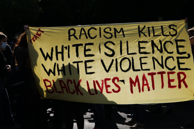 Eine Gruppe maskierter Personen mit einem Banner, auf dem "Rassismus tötet Weiße Stille, weiße Gewalt, Black Lives Matter" steht, auf einer dunklen Straße.