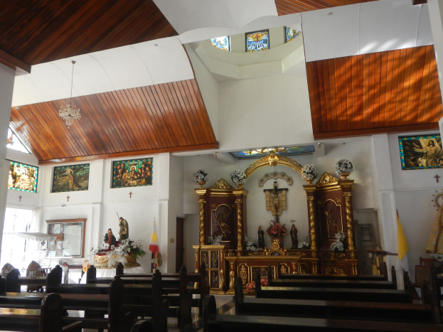 Innere eines Kirchenschiffs mit hölzernen Kirchenbänken, Statuen, Blumenbouquets, Fahnen, Fotorahmen an den Wänden und Buntglasfenstern, Decke mit Lichtern geschmückt.
