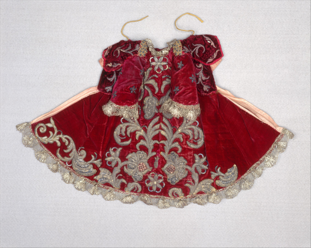 Ein rotes Samtkleid mit goldener Stickerei auf einem Bügel vor einem weißen Hintergrund.