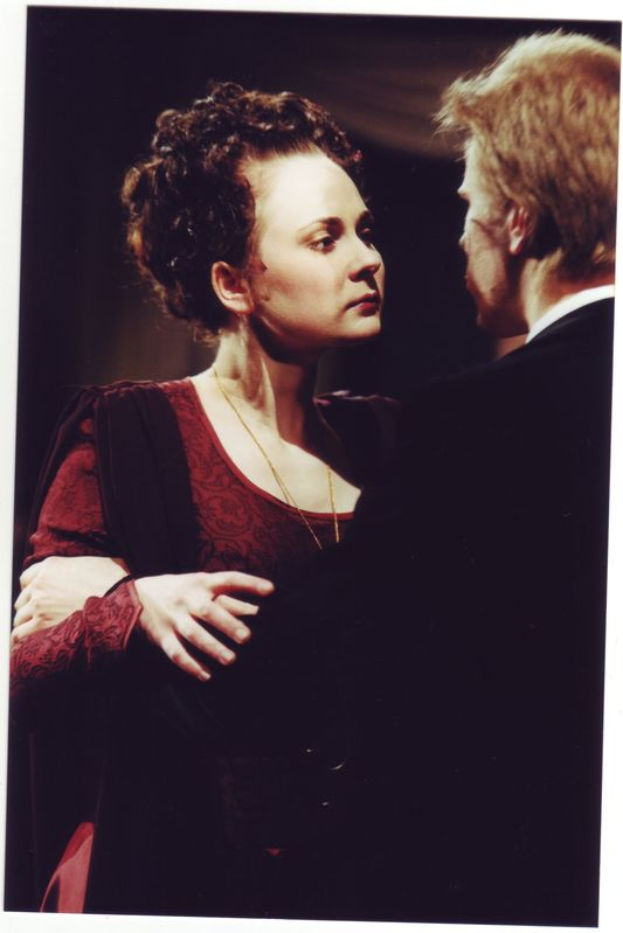 Eine Frau in einem roten Kleid und ein Mann in einem schwarzen Anzug stehen Seite an Seite, wobei der Mann die Frau mit liebevollem Blick ansieht, vor einem dunklen Hintergrund.