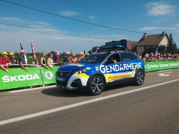 Polizeiauto fährt an einer Menge vorbei, die sich bei Bäumen und Gebäuden befindet, während eines Events mit Flaggen und einem Banner, auf dem "Gendarmerie de la Tour de France" steht.