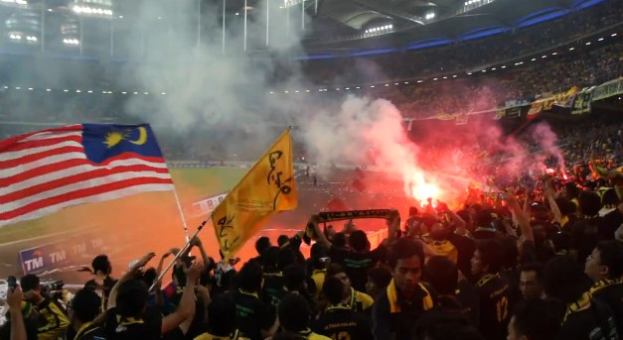 Eine große Menschenmenge in einem Stadion mit brennenden Bengalos in der Luft, einige halten Fahnen und eine Person hält eine Kamera, mit Rauch im Hintergrund und Schildern mit der Aufschrift "Malaysia vs Malaysia Live Streaming".