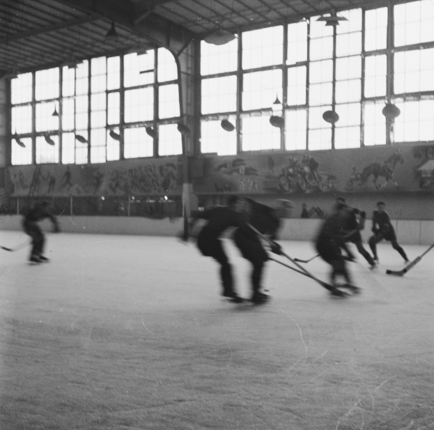 Schwarzes und weißes Foto von Menschen, die Hockey auf einem Eisplatz mit bemalten Wänden und Fenstern im Hintergrund spielen.
