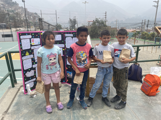 Gruppe von Kindern auf einem Basketballfeld, die Papiertüten halten, mit Tüten und Gegenständen rechts und einer Tafel mit Text links, vor einem Hintergrund aus Zaun, Pfählen, Straßenlaternen, Bäumen, Gebäuden, Hügeln und Himmel.