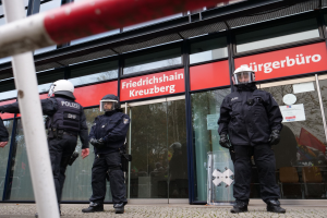 Polizeibeamte in Uniform vor einem gläsernen Gebäude mit angeklebten Papieren, einem Pfahl links und Hintergrundschildern mit der Aufschrift "Friedrichshain Kreuzberg."