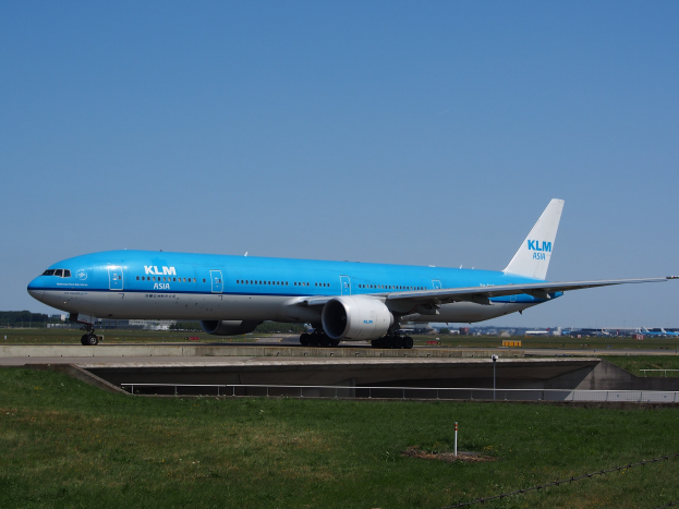 Ein KLM Boeing 777-300ER auf der Rollbahn am Amsterdam Airport, umgeben von Gras und einem Zaun mit Pfählen, mit Gebäuden, Bäumen und einem klaren blauen Himmel im Hintergrund.