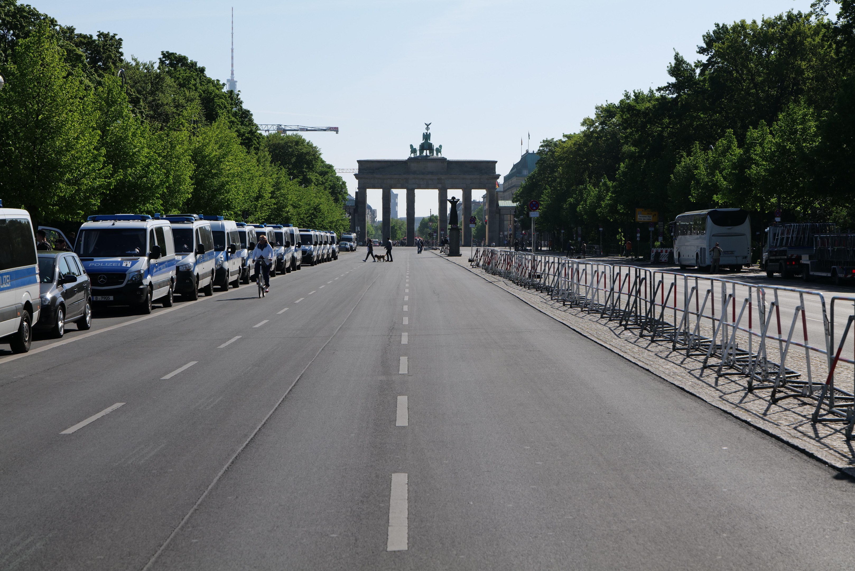 Lange Reihe von Polizeiwagen vor dem Brandenburger Tor geparkt, mit Fahrradfahrern, Fußgängern, Barrieren, Bäumen und einem Bogen mit Statuen im Hintergrund.