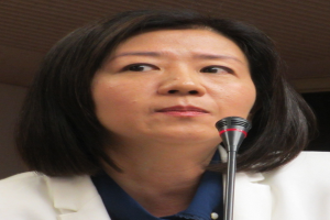 Frau in weißer Jacke, die in ein Mikrofon spricht, vor einer Wand stehend, mit ernstem Gesichtsausdruck