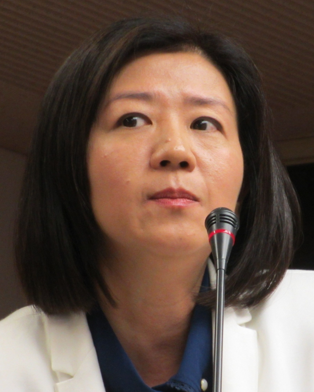 Frau in weißer Jacke, die in ein Mikrofon spricht, vor einer Wand stehend, mit ernstem Gesichtsausdruck