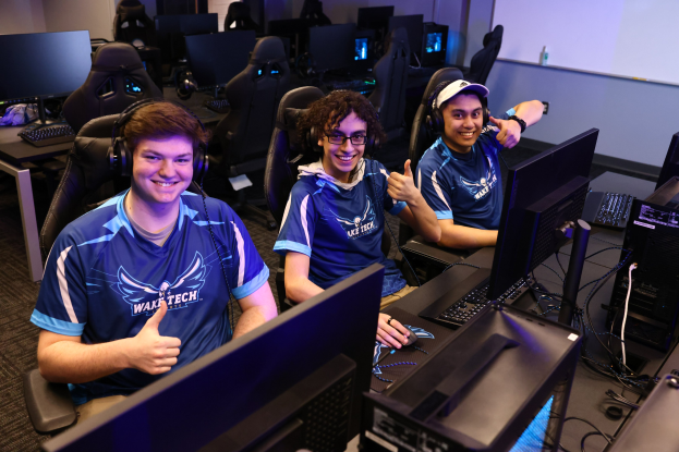 Drei junge Männer in Headsets lächeln, während sie an Computern während einer Esports-Gaming-Session sitzen, mit Monitoren, Tastaturen und Kabeln auf dem Schreibtisch und einem Bildschirm im Hintergrund.