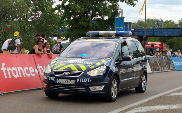 Polizeiauto fährt an einer Menschenmenge vorbei, die Schilder, Bäume, eine Brücke, eine Fahne und einen bewölkten Himmel im Hintergrund hat.