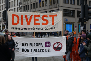 Gruppe von Menschen, die ein Banner mit der Aufschrift "Divest Hey Europe, Don't Frack Us" auf einer Straße mit Gebäuden, Straßeninfrastruktur, Fahrzeugen, Bäumen und einem klaren Himmel im Hintergrund halten.