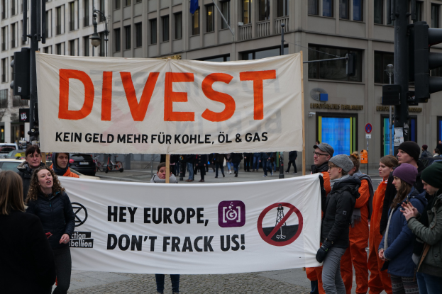 Gruppe von Menschen, die ein Banner mit der Aufschrift "Divest Hey Europe, Don't Frack Us" auf einer Straße mit Gebäuden, Straßeninfrastruktur, Fahrzeugen, Bäumen und einem klaren Himmel im Hintergrund halten.