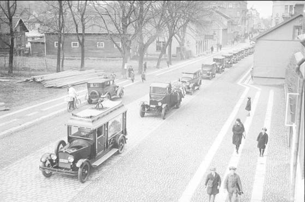 Schwarzes und weißes Foto einer Berliner Straße in den 1930er bis 1940er Jahren, das alte Autos, Fußgänger, Gebäude mit Fenstern, Bäume und einen klaren Himmel zeigt.