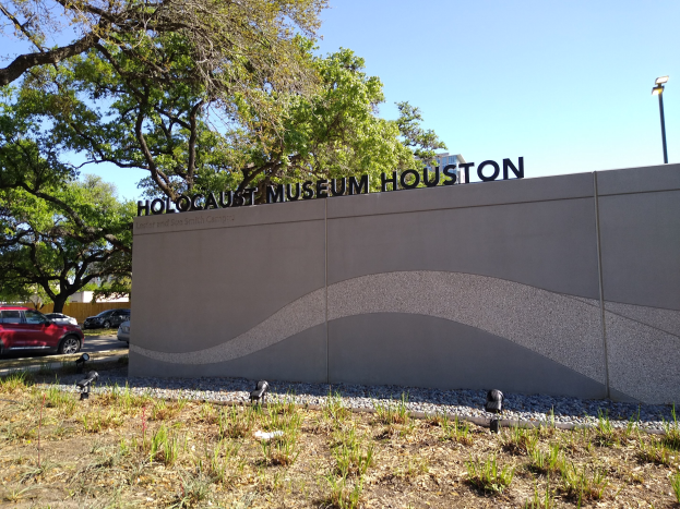 Außenansicht des Houston Holocaust Museums mit eingravierter Textwand, Gedenksteine, Grünfläche, Straße mit Fahrzeugen, einer Straßenlaterne und bewölktem Himmel.