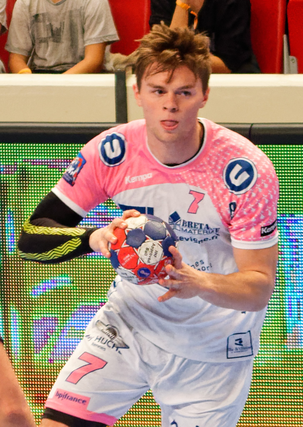 Ein Mann in einem pinken Hemd mit einem Fußball, mit einer anderen Person Hand auf der linken Seite, vor einem Hintergrund mit Menschen auf Stühlen sitzend.