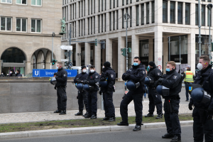 Polizisten in schwarzen Uniformen und Masken vor einem Berliner Gebäude mit Glasfenstern und Säulen, einige halten Helme, mit Laternen, Verkehrsampeln, Schildern mit Text, einer Statue und anderen Menschen im Hintergrund.