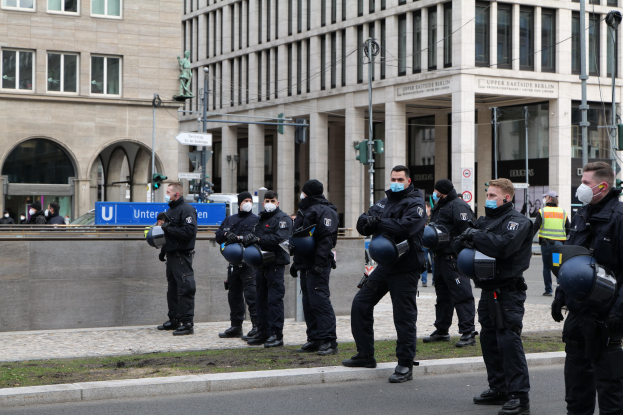 Polizisten in schwarzen Uniformen und Masken vor einem Berliner Gebäude mit Glasfenstern und Säulen, einige halten Helme, mit Laternen, Verkehrsampeln, Schildern mit Text, einer Statue und anderen Menschen im Hintergrund.