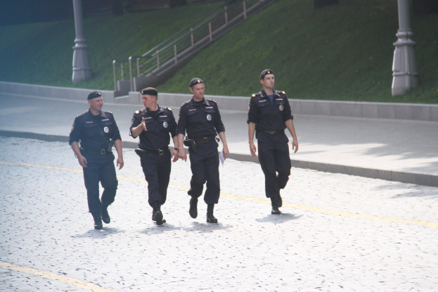 Drei Polizeibeamte in Uniform und Mützen, die eine Straße mit Geländern, Treppen und Gras im Hintergrund entlanggehen.