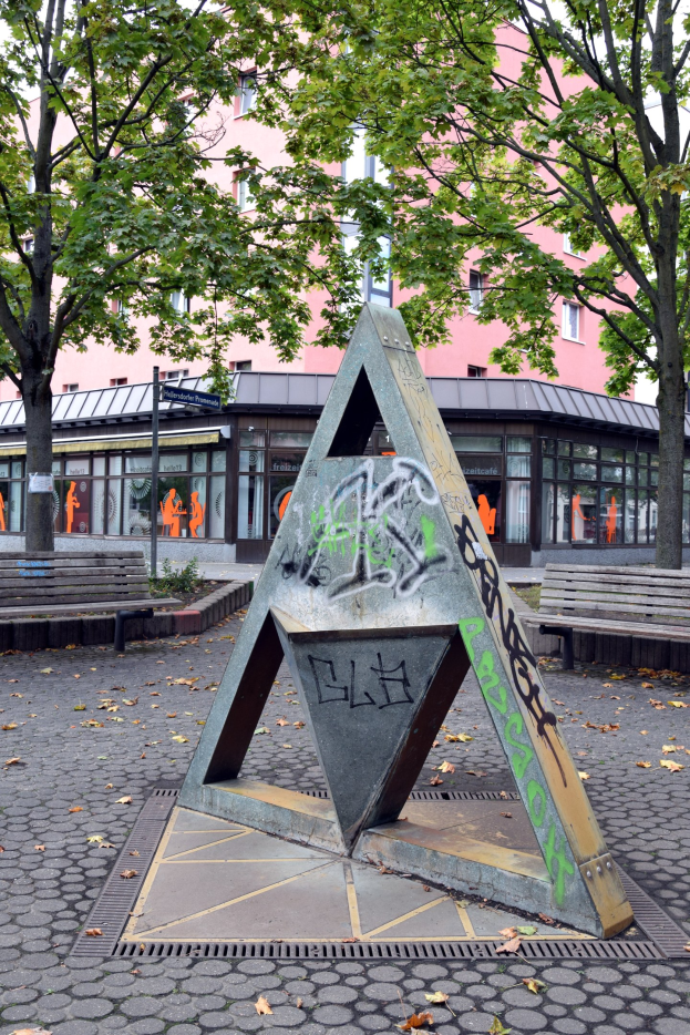 Metallskulptur mit Graffiti in einem Park, umgeben von Bäumen, Bänken, einem Pfahl und einem Gebäude im Hintergrund.