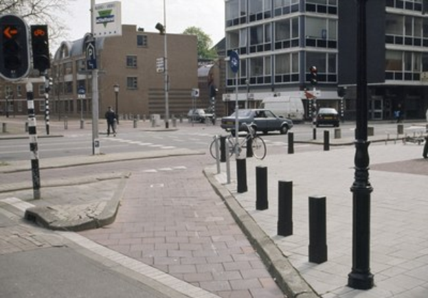 Eine Stadtstraße mit einem geparkten Fahrrad, fahrenden Fahrzeugen, Fußgängern, Versorgungsmasten, Verkehrsampeln, Schildern, Bäumen, Gebäuden mit Fenstern und einem klaren blauen Himmel.