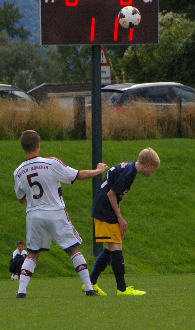 Zwei junge Männer beim Fußballspielen auf einem Rasenplatz mit Bäumen, Pflanzen und parkenden Autos im Hintergrund, mit einer nummerierten Tafel an einem Pfahl.