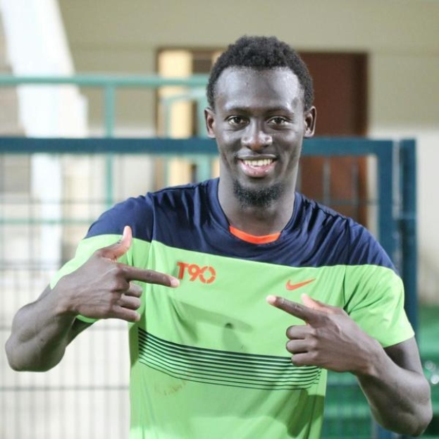 Ein lächelnder Mann in einem grünen und blauen Hemd zeigt auf die Kamera, mit einem unscharfen Geländer, Wand und Tür im Hintergrund.