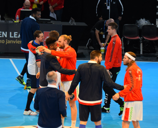 Mannengruppe auf einem Futsal-Platz mit Bällen, feiernd mit Stühlen, einem Tisch mit Flaschen und Laptops sowie einer Tafel mit Text im Hintergrund.