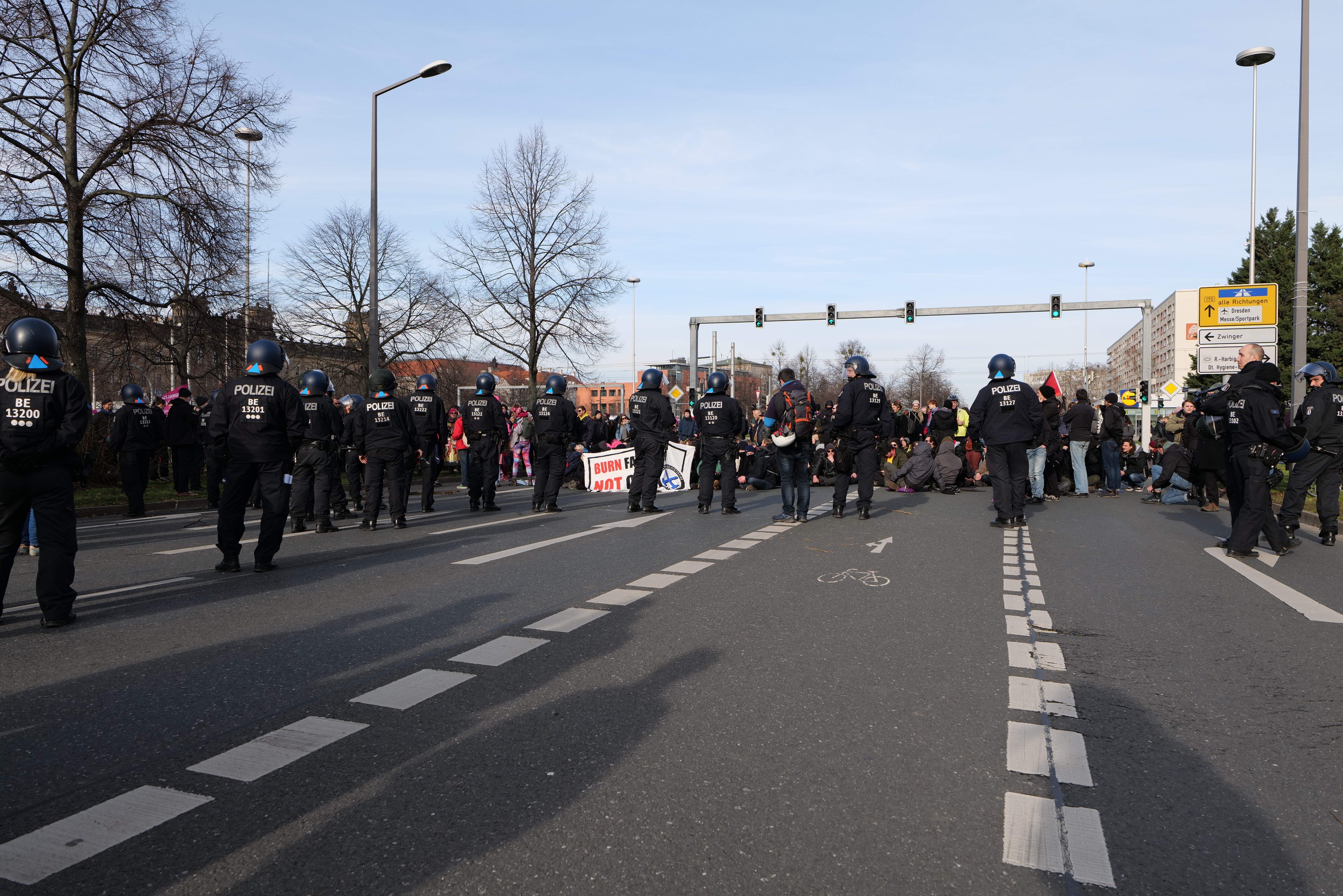 Eine Gruppe von Polizisten in schwarzen Uniformen und Helmen, die an der Straße mit Laternenmasten, Ampeln, Bäumen, Gebäuden und einem klaren blauen Himmel im Hintergrund stehen.