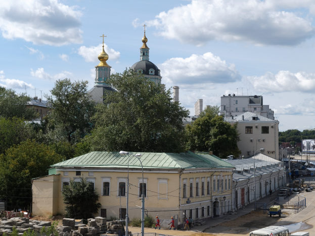 Ein Blick auf eine Stadt mit der Kirche der Versöhnung am Nerl im Hintergrund, umgeben von Gebäuden, Bäumen, Straßenmasten, Straßenlaternen, Strommasten, Stromkabel, Kraftfahrzeugen auf der Straße, Menschen, die auf dem Gehweg gehen, und Felsen unter einem bewölkten Himmel.