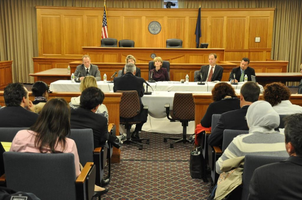 Ein Sitzungssaal mit einer Gruppe von Menschen, die auf Stühlen sitzen, zwei ausländische Flaggen, ein oranger Schreibtisch mit Wasserflaschen und einem Mikrofon.
