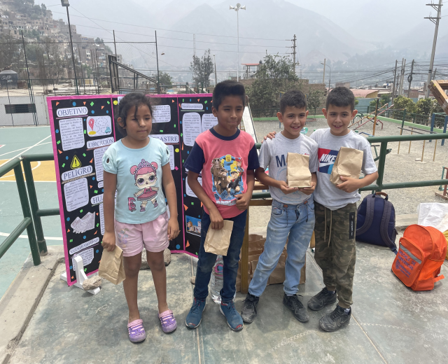 Gruppe von Kindern auf einem Basketballfeld mit Papierbeuteln in den Händen, mit Beuteln und Gegenständen rechts und einer Tafel mit Text links, vor einem Hintergrund aus Zaun, Pfosten, Straßenlaternen, Bäumen, Gebäuden, Hügeln und Himmel.